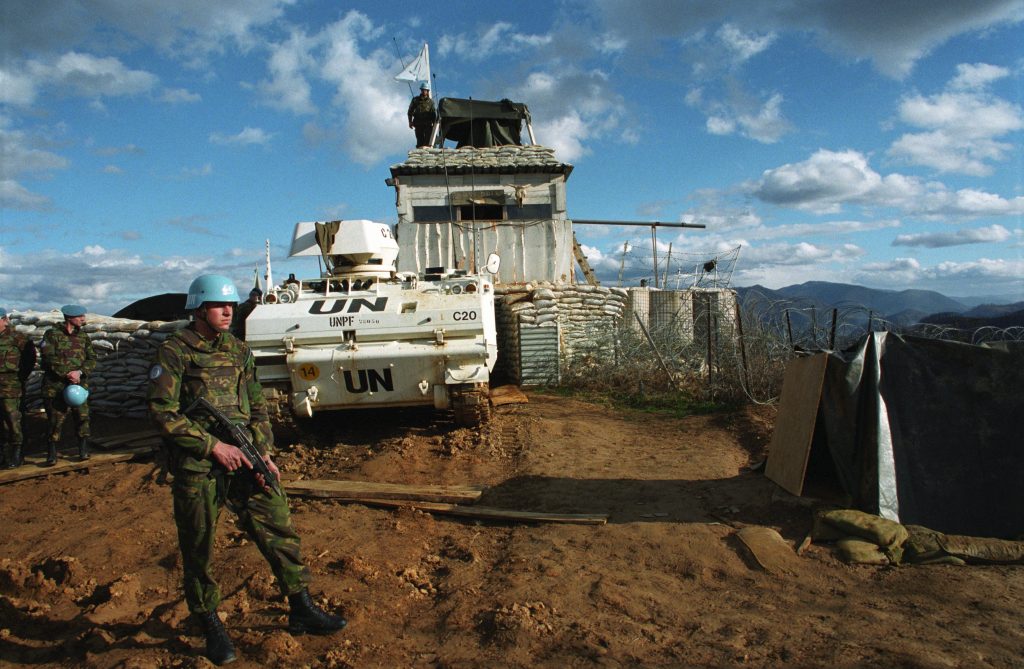 VN-militairen in camouflage-uniform met blauwe helmen staan bij een gepantserd voertuig met UN-markering, voor een geïmproviseerde post met zandzakken en wachttoren in bergachtig gebied.