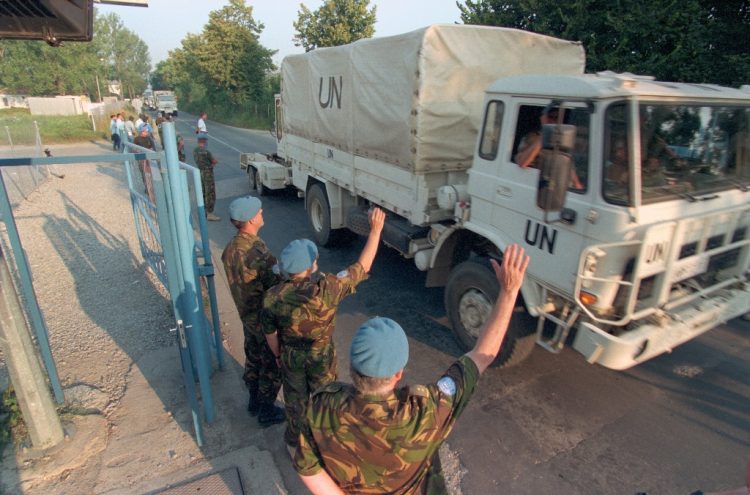 Drie VN-militairen in camouflage-uniform en blauwe helmen staan langs de weg en steken hun hand op naar een voorbijrijdende UN-vrachtwagen met huif. In de verte staan enkele burgers langs de weg te kijken.