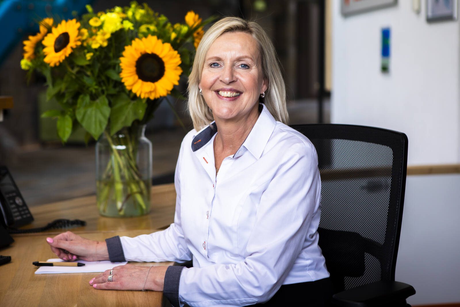 Vrouw zit glimlachend aan een bureau in een kantooromgeving, met een vaas zonnebloemen op de achtergrond.