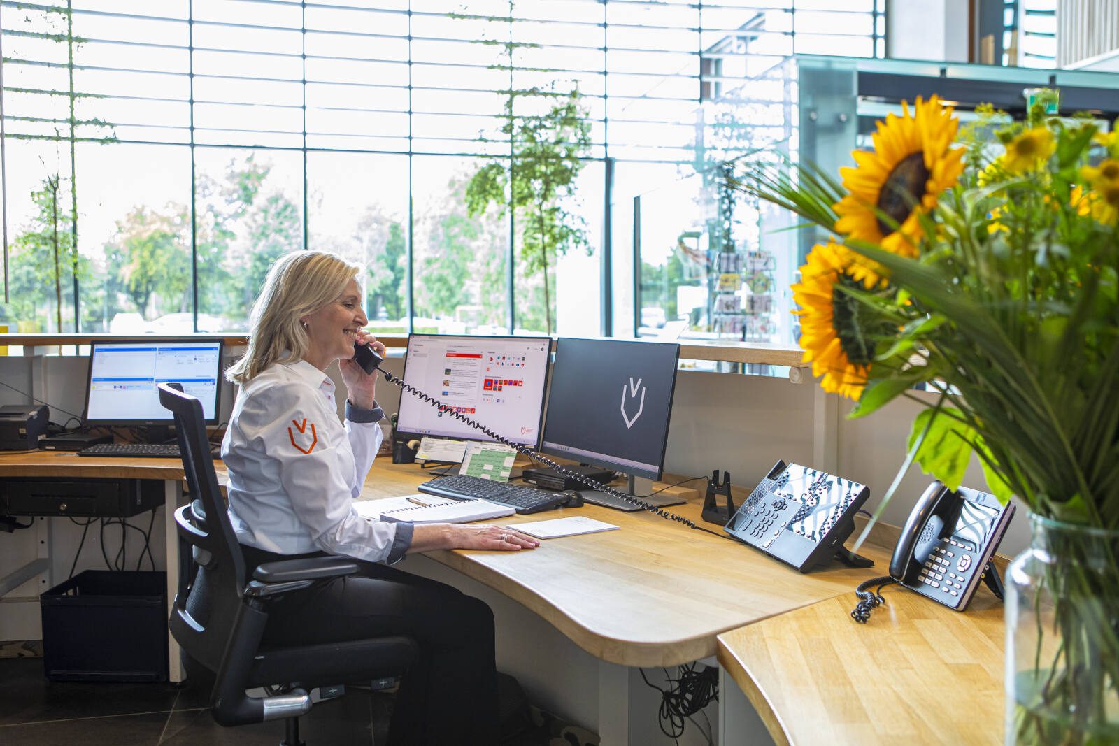 Vrouw zit aan een bureau in een lichte kantoorruimte en voert een telefoongesprek via een vaste telefoon. Voor haar staan meerdere computerschermen met open programma’s. Op de voorgrond staat een vaas met zonnebloemen, en achter haar zijn grote ramen met uitzicht op groen.