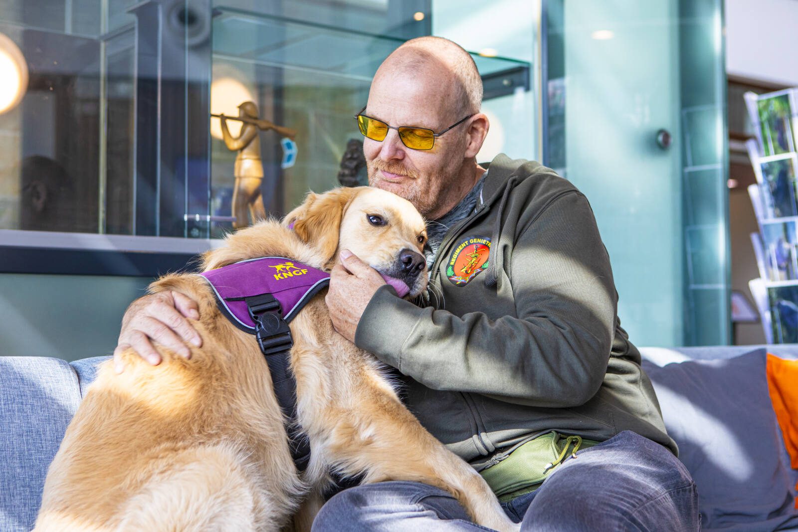 Kale man met gele bril zit op een bank en knuffelt een golden retriever met een paars hulphondenharnas, in een moderne binnenruimte met glazen wanden op de achtergrond.