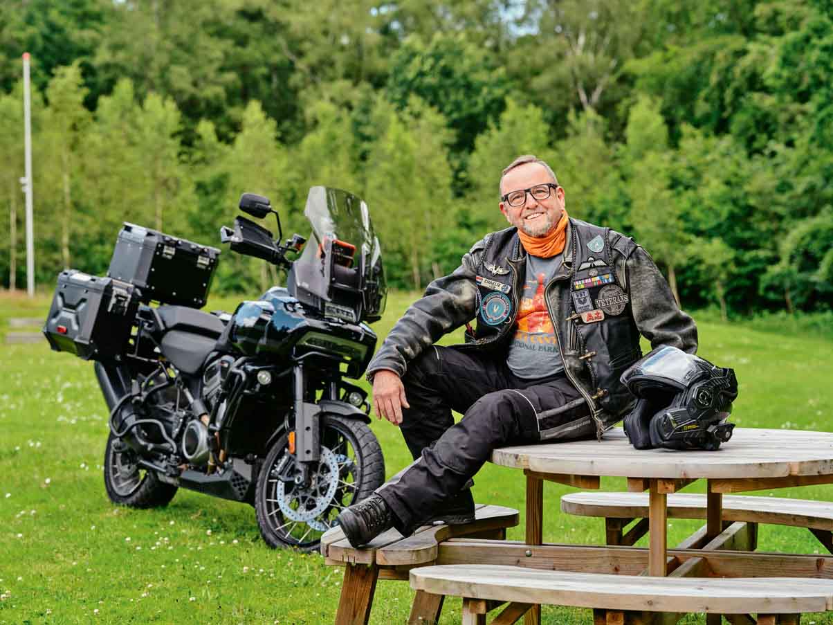 Collega Charlie Malschaert in motorkleding met diverse emblemen zit ontspannen op een picknicktafel in een groen park. Naast hem staat een grote toermotor met koffers, en op de tafel ligt zijn motorhelm.