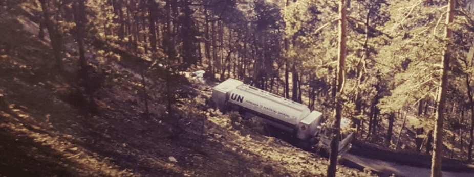 Een UN-tankwagen ligt schuin op een steile, beboste helling tussen hoge bomen, met zonlicht dat door het bos valt.