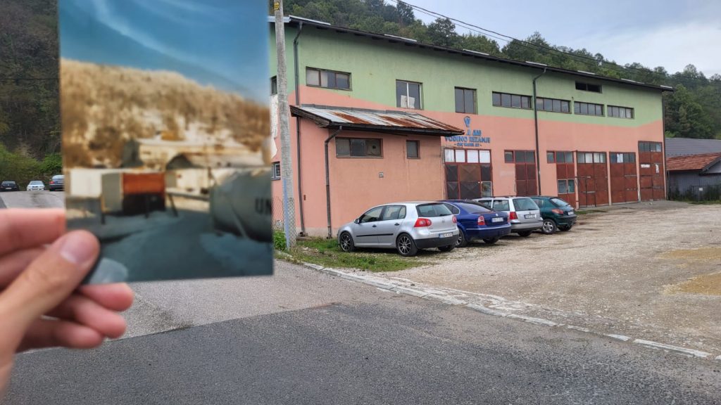 Een hand houdt een oude, enigszins vervaagde foto omhoog van een gebouw met UN-markering en tanks, terwijl op de achtergrond op dezelfde locatie een huidige fabriekshal met groen-roze gevel en geparkeerde auto’s te zien is.