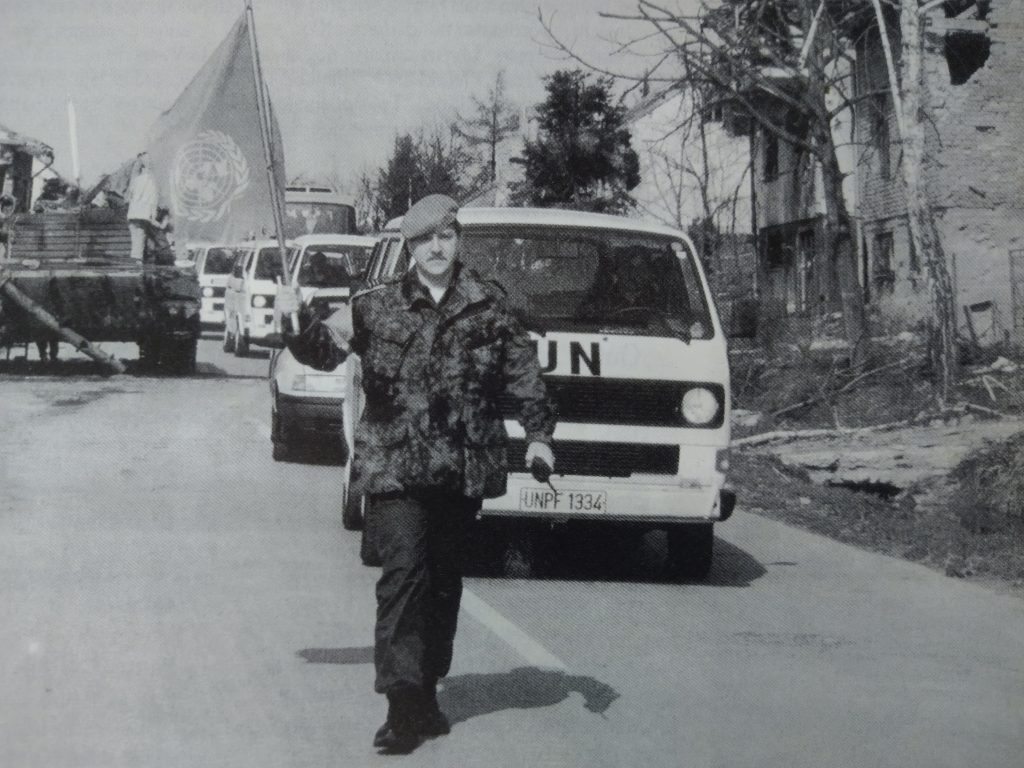 Zwart-witfoto van een VN-militair met baret die voorop loopt met een VN-vlag, gevolgd door een colonne voertuigen met UN-markering in een beschadigde straat.