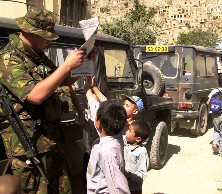 Een militair in camouflage-uniform leest een document terwijl enkele kinderen naast hem nieuwsgierig meekijken en proberen het papier aan te raken. Op de achtergrond staan terreinwagens geparkeerd in een stoffige straat met bebouwing tegen een heuvel.