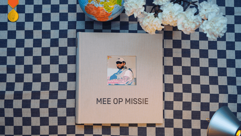 Bovenaanzicht van een boek met de titel “MEE OP MISSIE” op een blauw-wit geruite tafelkleed. Op de kaft staat een illustratie van een militair; daarnaast liggen een wereldbol, witte bloemen en een medaille.