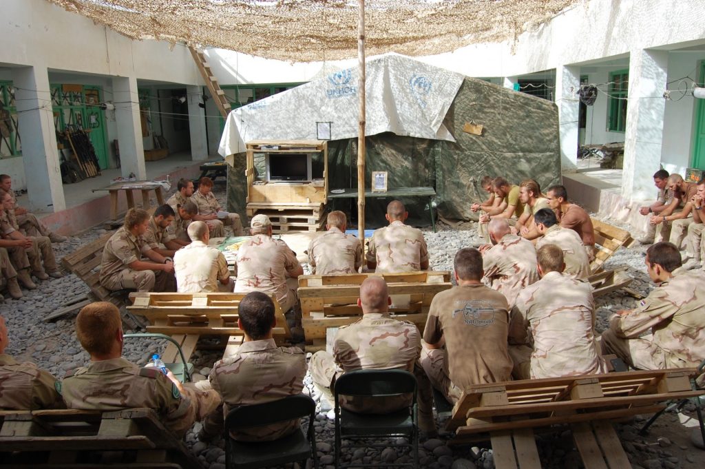 Groep militairen in camouflage-uniform zit op houten banken in een binnenplaats rond een tent met VN-logo, terwijl zij gezamenlijk luisteren of overleggen in een geïmproviseerde bijeenkomst.