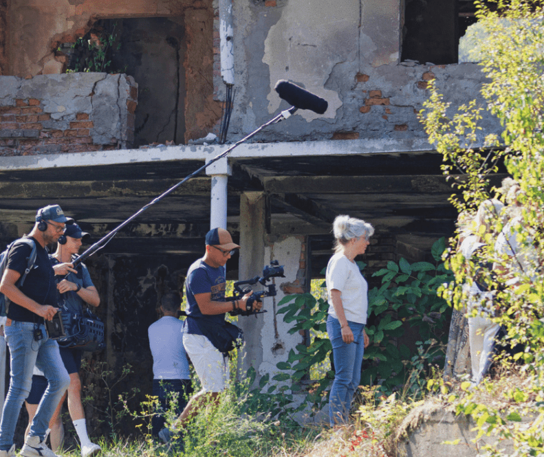 Filmcrew met geluidsman en cameraman filmt een vrouw bij een beschadigd en deels verwoest gebouw in Srebrenica; op de achtergrond zijn afbrokkelende muren en begroeiing zichtbaar.
