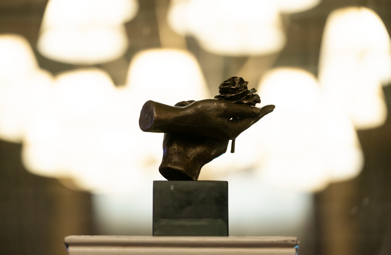 Bronzen beeldje van een hand met een witte anjer op een sokkel, met op de achtergrond onscherpe lichten in een zaal.