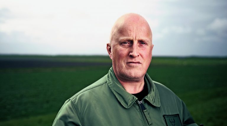 Portret van een kale man in een groene jas die recht in de camera kijkt, met op de achtergrond een uitgestrekt groen landschap onder een bewolkte lucht.