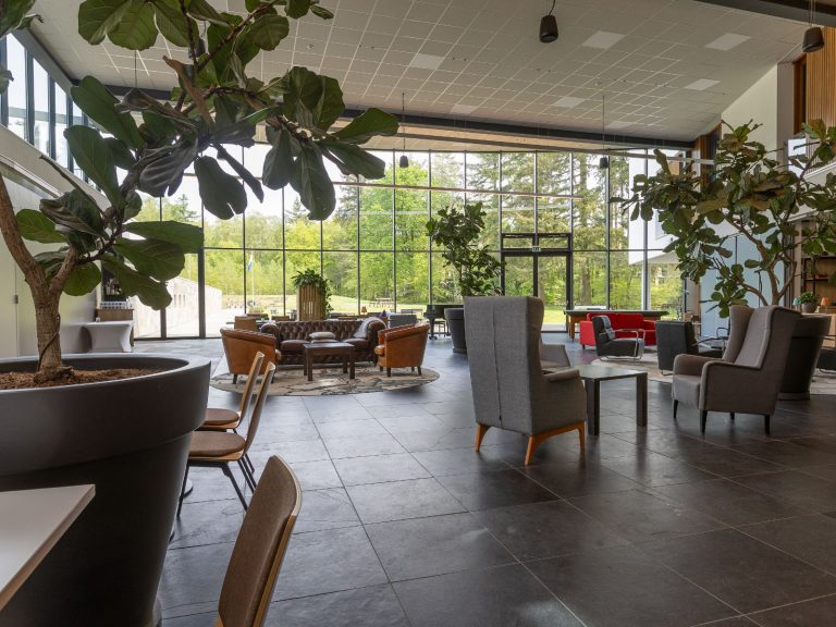 Ruime lobby met zitgedeelte, grote planten en hoge ramen met uitzicht op bomen.