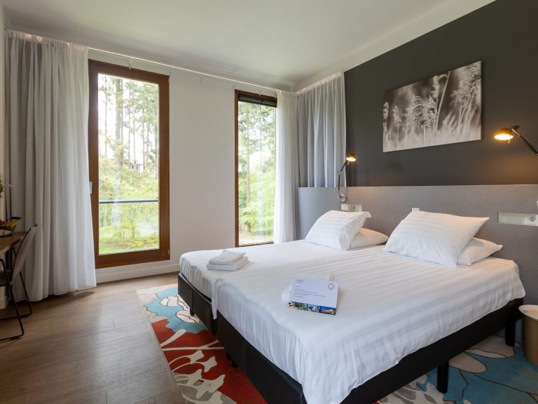 Moderne hotelkamer met tweepersoonsbed met wit beddengoed en opgevouwen handdoeken. Aan weerszijden van het bed hangen leeslampjes aan een donkere accentmuur met een zwart-wit kunstwerk erboven. Grote ramen met witte gordijnen bieden uitzicht op bomen en groen. Op de vloer ligt een kleurrijk vloerkleed.