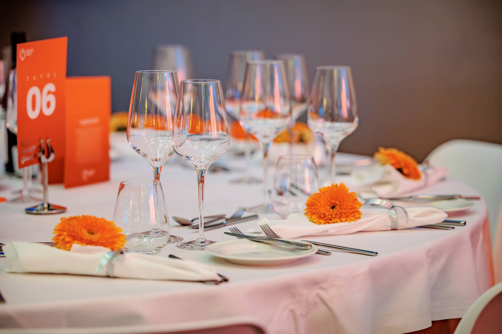 Gedekte tafel met wijnglazen, bestek en oranje bloemen tijdens een evenement.