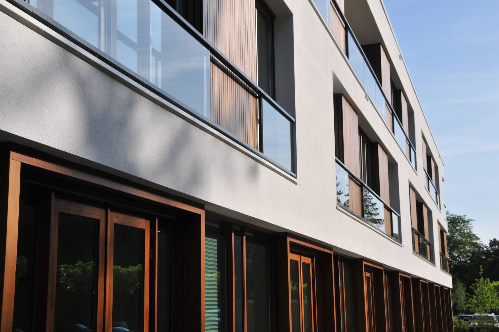 Gevel van een modern gebouw met witte muren, grote ramen en houten kozijnen, gezien vanuit een schuin perspectief.