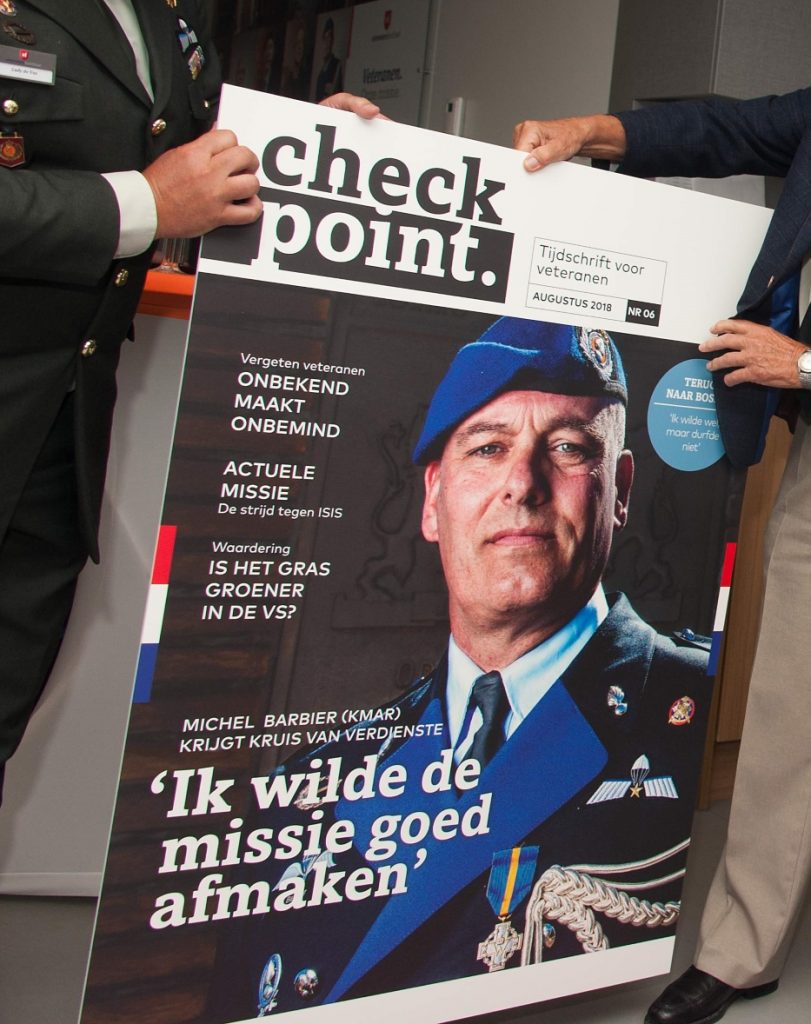 Twee mannen houden samen een groot bord vast met de cover van het tijdschrift “Check Point”, waarop een militair in ceremonieel uniform met blauwe baret en onderscheidingen staat afgebeeld, omringd door koppen over veteranen en missies.