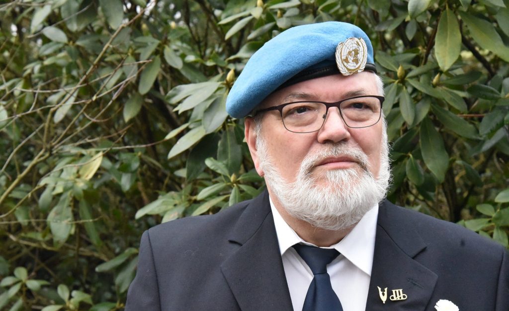 Portret van een oudere man met bril en witte baard, gekleed in een donker pak met stropdas en een blauwe VN-baret met embleem, staand voor een achtergrond van groene struiken.