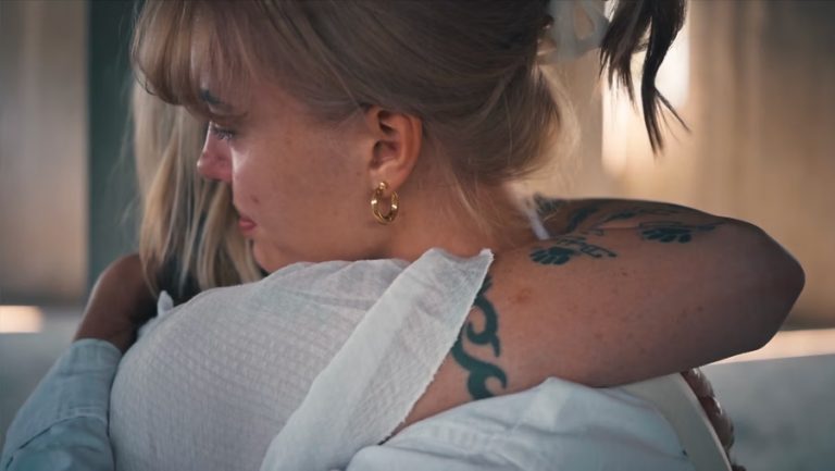 Close-up van twee vrouwen die elkaar omhelzen; één vrouw met een staart en gouden oorbellen kijkt emotioneel opzij, terwijl de andere vrouw haar arm met een zichtbare tatoeage om haar heen slaat.
