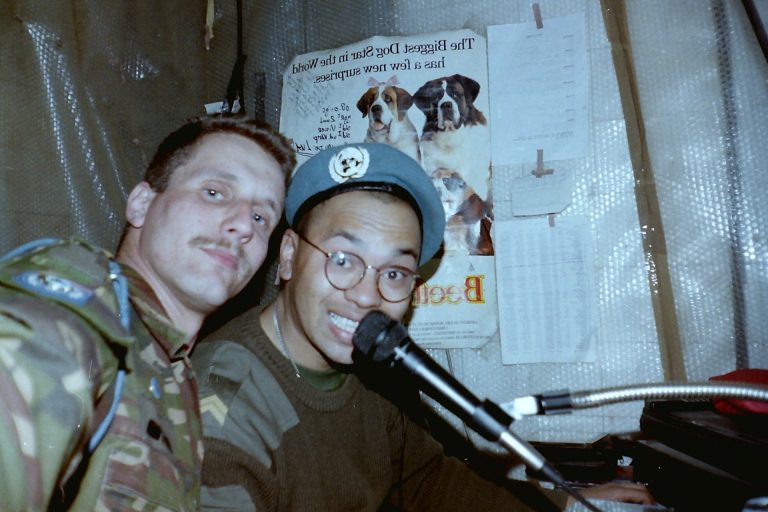 Twee militairen poseren in een geïmproviseerde radiopost; één in camouflage-uniform kijkt in de camera, terwijl de ander met bril en blauwe baret in een microfoon spreekt. Op de achtergrond hangen papieren en een poster aan de wand.