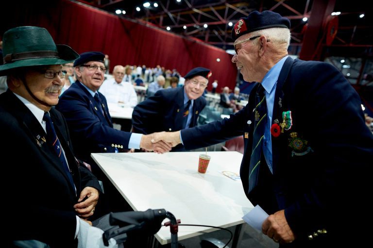Twee oudere mannen in uniformachtige kleding en baretten schudden elkaar de hand aan een tafel tijdens een bijeenkomst. Beiden dragen onderscheidingen en poppies op hun jasje. Op de achtergrond zitten andere mannen in vergelijkbare kleding in een grote zaal met rode wanden.