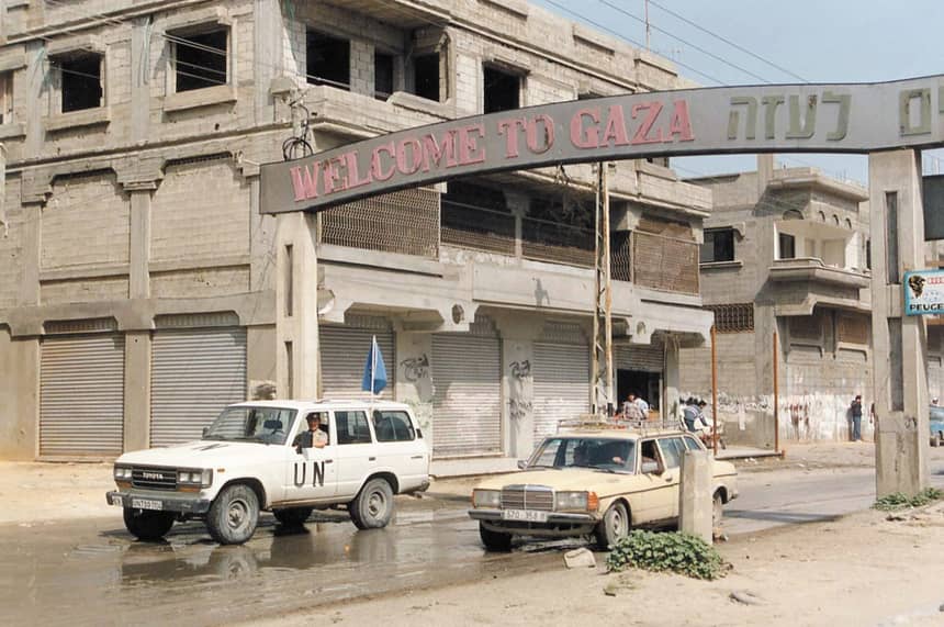 Een straatbeeld in Gaza met een poort waarop “Welcome to Gaza” staat. Op de voorgrond rijden een witte VN-terreinwagen en een personenauto over een natte weg. Op de achtergrond staan beschadigde gebouwen met gesloten rolluiken en enkele mensen op straat.