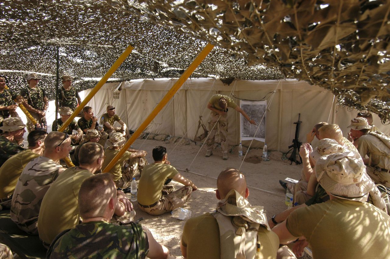 Militairen zitten onder een camouflagenet terwijl een instructeur bij een kaart een briefing geeft op een zandachtige buitenlocatie.