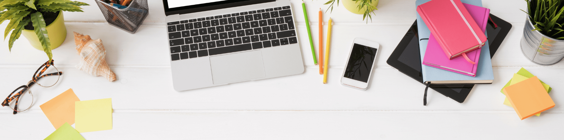 Bovenaanzicht van een opgeruimd bureau met een laptop in het midden, omringd door kleurrijke notitieboekjes, potloden, een smartphone en post-its. Aan de zijkanten liggen een bril, een schelp en enkele planten, wat zorgt voor een frisse en creatieve werkplek.