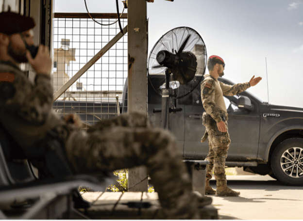 Twee militairen in camouflage-uniform en rode baretten bevinden zich bij een controlepost. Op de voorgrond zit een militair ontspannen op een stoel, terwijl een ander naast een voertuig staat en met zijn arm een richting aanwijst. Op de achtergrond zijn een ventilator, hekwerk en een pick-uptruck zichtbaar.