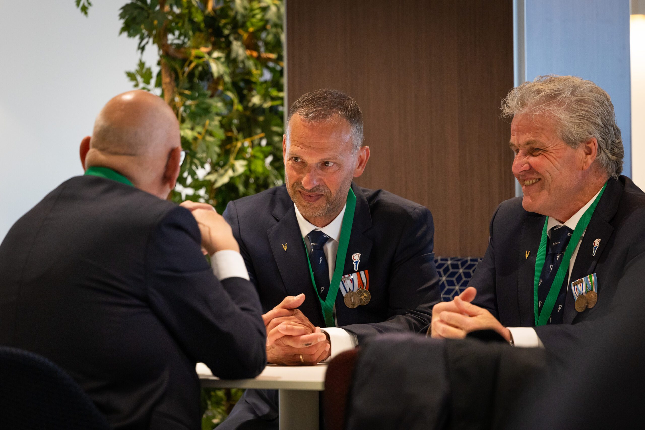 Drie veteranen in nette pakken zitten aan een tafel in gesprek. Twee van hen dragen medailles en groene linten om hun nek en lachen terwijl ze aandachtig luisteren naar de man met zijn rug naar de camera. De setting oogt als een formele bijeenkomst, met een plant en houten wand op de achtergrond.