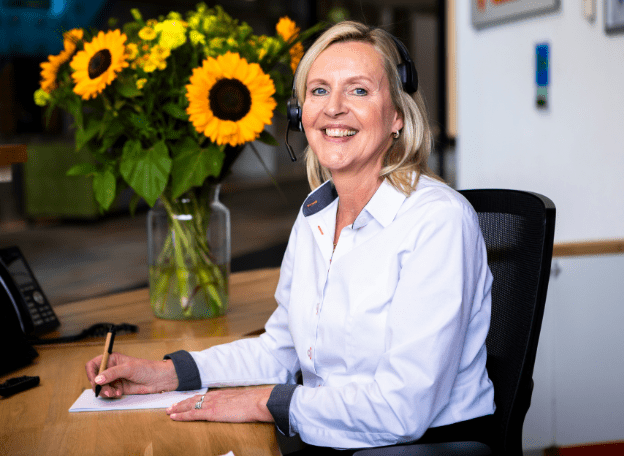 Vriendelijke receptioniste met headset zit aan een bureau en schrijft iets op een notitieblok, met een vaas zonnebloemen op de achtergrond in een kantooromgeving.