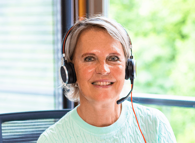 Medewerker Veteranenloket met kort blond-grijs haar draagt een headset en glimlacht terwijl ze achter een bureau bij een raam zit, met groen landschap op de achtergrond.
