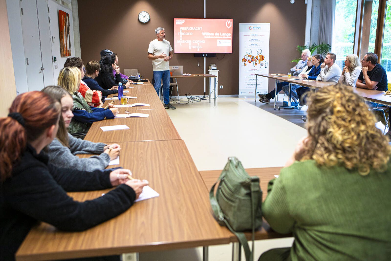 Trainer geeft een presentatie over veerkracht aan een groep deelnemers die aan lange tafels in een vergaderruimte zitten en aandachtig luisteren naar het scherm aan de voorkant.