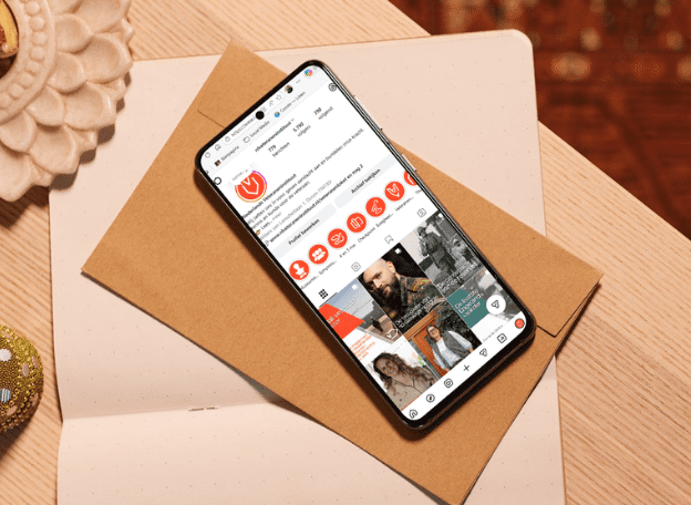 Een smartphone ligt schuin op een bruine envelop op een lichte houten tafel. Op het scherm is een Instagram-profiel geopend met een wit en oranje kleurenschema en meerdere ronde iconen en fotoposts zichtbaar. Links bovenin ligt een decoratief schaaltje en rechtsonder een klein glinsterend object.