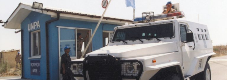 wit VN‑voertuig met een militair staant uit het dakraam en blauwe controlepost van missie UNFICYP bij een poort in de bufferzone op Cyprus.