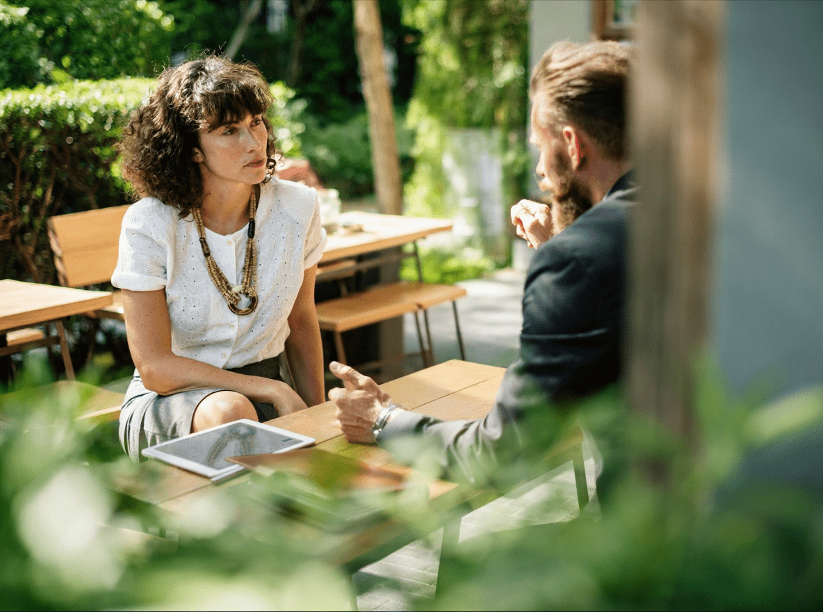 Twee mensen in gesprek aan een tafel buiten; een vrouw luistert aandachtig terwijl een man iets toelicht met een laptop en tablet op tafel.