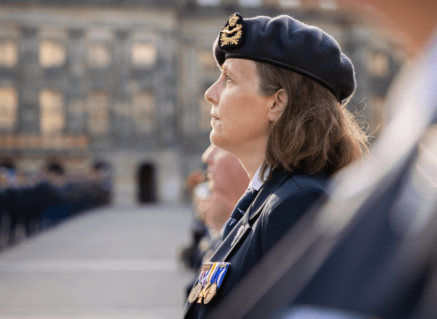 Zijaanzicht van een vrouw in uniform met een donkere baret en onderscheidingen op haar borst, die geconcentreerd voor zich uit kijkt tijdens een ceremonie. Op de achtergrond zijn andere deelnemers en een historisch gebouw zichtbaar, licht onscherp.