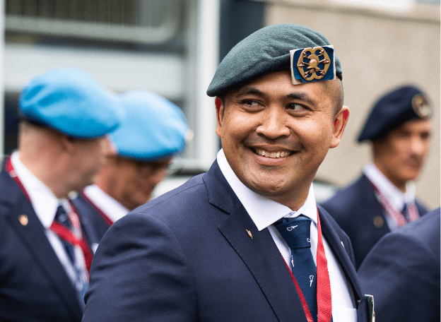 Een man in een net pak en groene baret kijkt glimlachend opzij tijdens een oefening voor de erecouloir. Op de achtergrond staan andere mannen in vergelijkbare kleding en verschillende baretten, licht onscherp in beeld.