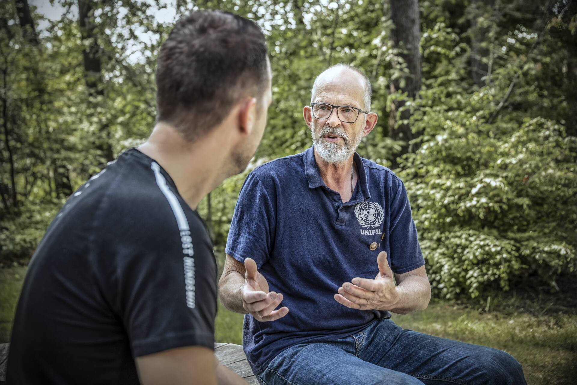 Twee mannen zitten buiten in een groene omgeving op een bankje en voeren een gesprek, waarbij een oudere man met een UNIFIL-shirt iets uitlegt.