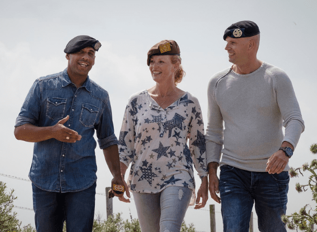 Drie veteranen lopen naast elkaar in de buitenlucht en praten ontspannen; ze dragen baretten en casual kleding en lijken in gesprek met elkaar.