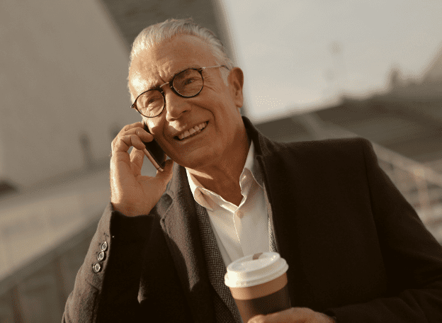 Een glimlachende oudere man met een bril praat buiten op zijn mobiele telefoon terwijl hij een koffiebeker vasthoudt. Hij draagt een nette jas en staat in warm zonlicht, met een onscherpe stedelijke achtergrond.
