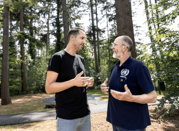 Twee mannen staan in een bosrijke omgeving en zijn met elkaar in gesprek. Ze maken gebaren met hun handen terwijl ze tegenover elkaar staan op een zonnig pad, omringd door hoge bomen.