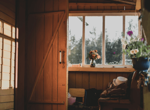 Rustieke houten schuur of kamer met een open deur en zonlicht dat naar binnen valt, een raam met uitzicht op groen buiten, en een vensterbank met bloemen in vazen; binnen staan een bankje met kleding en een hoed, wat zorgt voor een warme, landelijke sfeer.