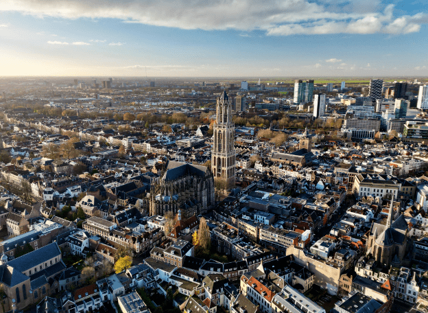 Luchtfoto van de binnenstad van Utrecht met de Domtoren centraal in beeld, omringd door historische gebouwen, grachten en straten, met moderne hoogbouw in de verte onder een deels bewolkte lucht.
