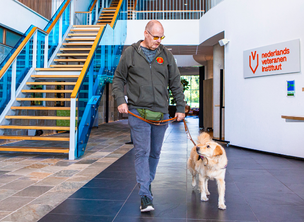 Een man met een bril en casual kleding loopt door een modern gebouw terwijl hij een hond aan de lijn heeft. De hond kijkt op naar de man terwijl ze samen over een tegelvloer lopen. Op de achtergrond is een open ruimte met een trap met blauwe leuningen en een muur met een logo en de tekst “nederlands veteranen instituut”.