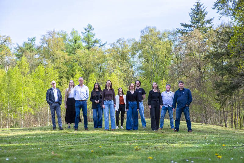 Een groep van elf mensen staat naast elkaar op een grasveld, omringd door groene bomen en struiken. De groep kijkt in de camera en poseert ontspannen in een natuurlijke, zonnige buitenomgeving.