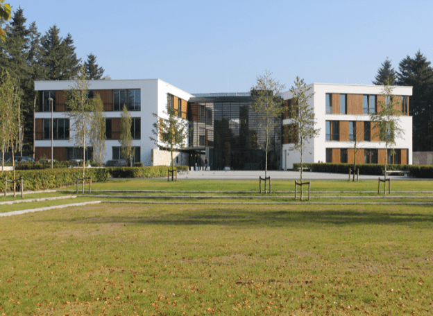 Een modern wit gebouw staat aan een groot grasveld met jonge bomen. De architectuur en de groene omgeving geven het terrein een open en rustige uitstraling.