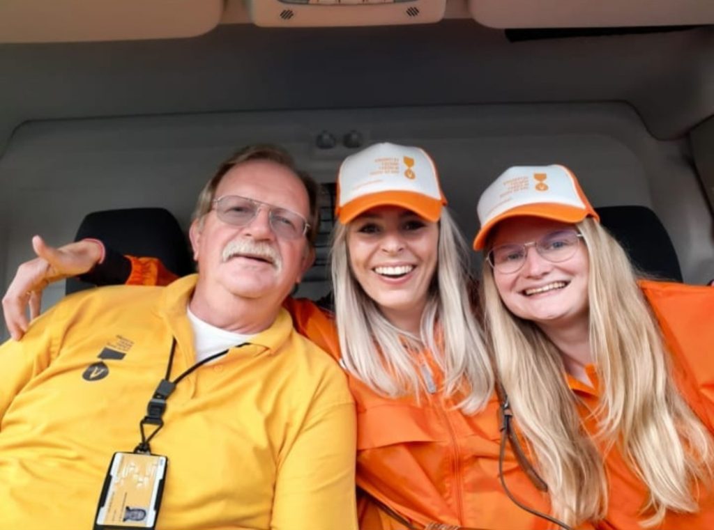 Drie vrijwilligers zitten lachend naast elkaar in een voertuig. Twee vrouwen dragen oranje petten en oranje werkkleding, terwijl de man links een geel shirt draagt. Ze kijken vrolijk in de camera