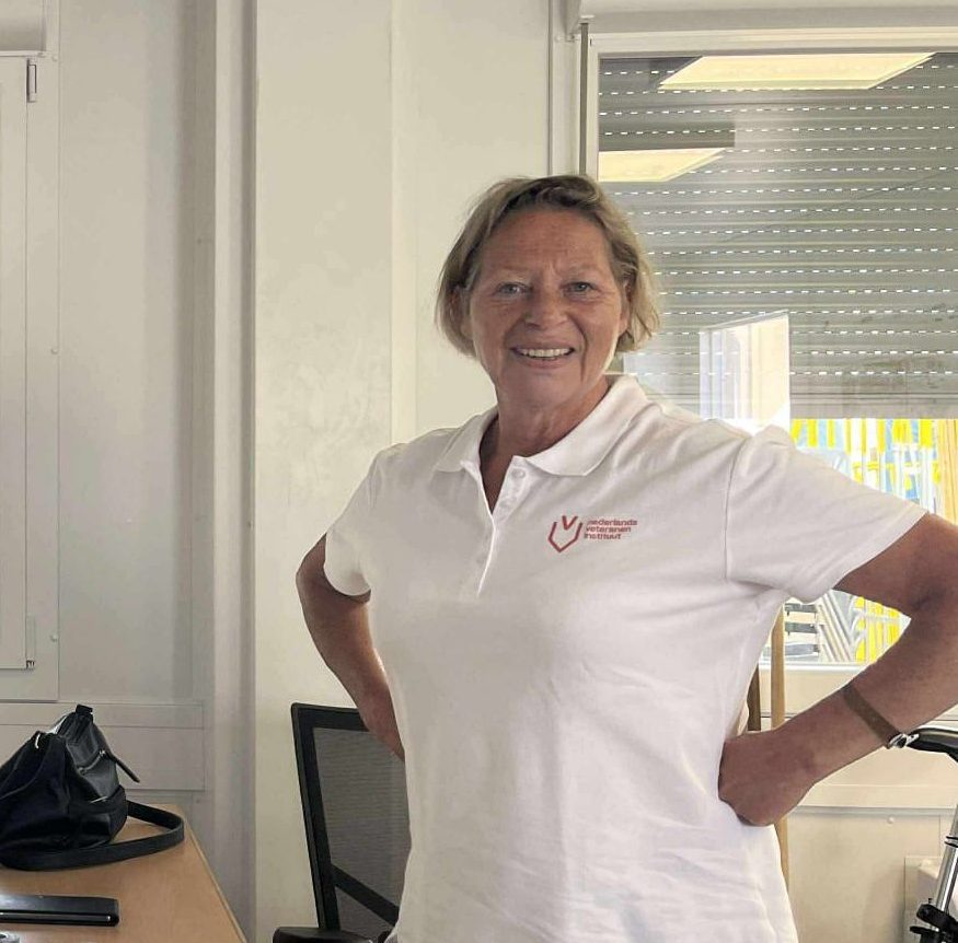 Een glimlachende persoon staat in een lichte kantoorruimte met de handen in de zij. De persoon draagt een wit poloshirt met een rood logo op de borst. Op de achtergrond zijn een bureau, een bureaustoel en grote ramen met halfgesloten zonwering zichtbaar.