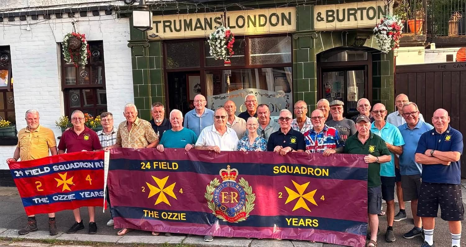 Groep van ruim twintig veteranen en oud militairen poseert voor The Cannon pub in Brompton. De deelnemers staan op de stoep voor de historische pub en houden twee vaandels van 24 Field Squadron Royal Engineers vast. De bijeenkomst weerspiegelt de sterke band tussen voormalige militairen en de traditie van reünies en kameraadschap die kenmerkend is voor deze veteranenvriendelijke ontmoetingsplaats. De gevel van de pub met haar groene tegels, bloemenmanden en historische bierreclames vormt de achtergrond van de groepsfoto.