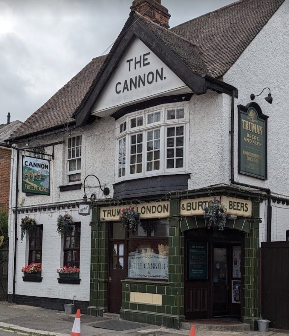 Gevel van café The Cannon, een traditionele Engelse pub met witte muren, donkergroene tegels rond de ingang en hangende bloembakken. Op de voorgevel staan verschillende borden met de naam van de pub en reclame voor bieren. De lucht is grijs en bewolkt, en voor het gebouw staan twee verkeerskegels op de stoep.